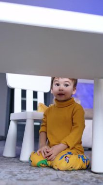 İki yaşında tatlı bir çocuk masanın yanında dizlerinin üstünde oturuyordu. Sarı elbiseli bebek yerde sürünüyor. Dikey video