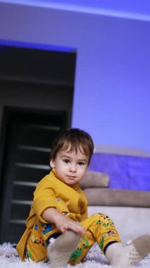 Koyu saçlı erkek bebek halının üzerinde oturuyor. Çocuk ekose kumaşla yatıp yuvarlanıyor. Dikey video