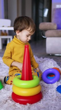 Huzurlu Kafkasyalı çocuk yerde yumuşak ekose üzerinde oturuyor. Şirin bebek piramidi birleştiriyor. Dikey video