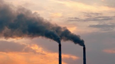 İki endüstriyel bacadan gün batımı gökyüzüne doğru duman yükseliyor. Arka planda güneş batarken iki uzun bacadan siyah duman yükseliyor..