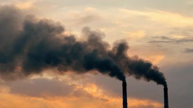 Dumanlar günbatımında endüstriyel bir alanda duman yayar. İki baca, gün batımında bir fabrikanın yakınında renkli bir gökyüzüne karşı yoğun duman üretiyor..