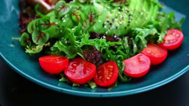 Masada mavi bir tabakta servis edilen domatesli ve yeşillikli taze salata. Bir tabakta taze sebzeler, dilimlenmiş domatesler ve diğer salata malzemeleri var..