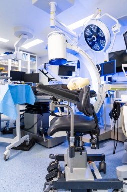 Mobil C-kolu x-ray makinesi ve ameliyathanede sandalye. C-kolu floroskopi sisteminin geniş dikey görüntüsü ve nöroşirürji odasında özel bir sandalye. Arkaplanda cerrahi ışıklar, monitörler ve instrume ile steril tablolar yer almaktadır.