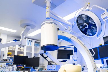 Cerrahi ışık ve beyin cerrahisinde C-kolu ayrıntıları. Düşük açılı çekim büyük dairesel cerrahi bir LED lambaya ve C-kolu görüntüleme sisteminin üst kısmına odaklanıyor. Klinik arka planda beyaz bir tavan.