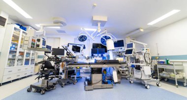 Gelişmiş sinir cerrahisi ameliyathanesini geniş açıdan görebiliyoruz. C-kolu, ameliyat ışıkları, birden fazla monitör, anestezi makinesi ve cerrahi masaların yer aldığı geniş açılı bir nöroşirürji odası. Temiz beyaz duvarlar ve mavi renkli ışık