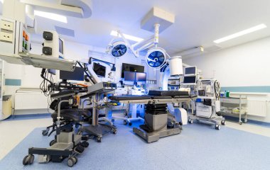 Cerrahi masa ve tıbbi görüntüleme cihazlarının düşük açılı görüntüsü. İç çekim, temiz bir klinikte robotik kollar, tıbbi monitörler ve özel cerrahi aydınlatma ile çevrili ayarlanabilir bir ameliyat masasına odaklanıyor..