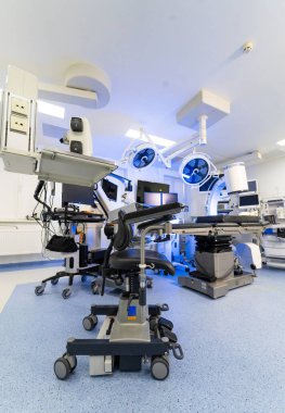 Yüksek teknolojili ameliyat masasının önünde ergonomik cerrah koltuğu. Görüntüleme sistemleri ve monitörlerle donatılmış modern bir ameliyathanede kollukları olan mobil cerrah taburesinin ön planı.