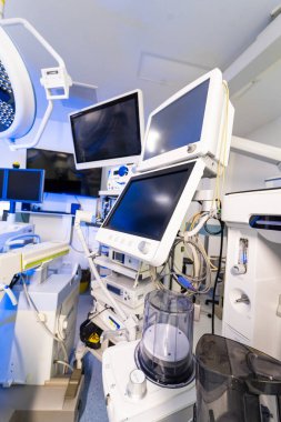 Klinikteki tıbbi izleme rafı ve anestezi ekipmanları. Cerrahi odadaki çoklu elektronik ekranların ve tıbbi cihazların dikey görüntüsü.