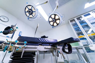 Steril hastane odasındaki ayarlanabilir ameliyat masasını kapatın. Parlak bir klinik ortamında bacak destekleri ve tıbbi dolapları olan mekanik bir ameliyat masasının ayrıntılı görüntüsü.