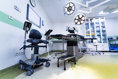 Ameliyat masası ve seyyar ergonomik koltuğu olan bir ameliyathane. İçinde cerrahi yatak, üstten lambalar ve özel yuvarlanan ergonomik sandalye bulunan bir tıbbi oda..