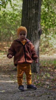 Sarı şapkalı, takım elbiseli ve ceketli sevimli çocuk parkta yürüyor. Küçük çocuk bir parmağıyla yukarıya doğru koşuyor. Dikey video