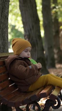 Parktaki bankta oturan tatlı çocuk. Bebek meyve püresi yer ve ayağını sallar. Dikey video