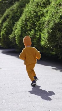 Sarı elbiseli ve şapkalı bir çocuğa yetişmek. Bebek parktaki ara sokağa doğru koşuyor. Dikey video