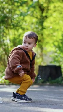 Beyaz çocuk kahverengi ceket giyip sabun köpüğü yakalıyor. Küçük oğlan parkta yürürken eğleniyor. Dikey video