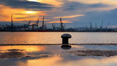 Büyük bir su birikintisinin yanındaki rıhtımda büyük bir demirleme bölmesi. Konteynır vinçlerinin silueti arka planda patlıyor. Gün batımında limana bak.