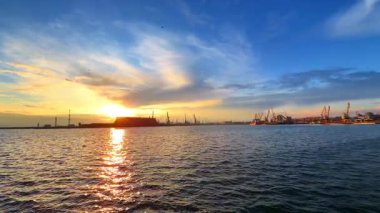 Batan güneşin ışığı su manzarasına yansıyor. Arkaplanda çok sayıda konteynır vinci bulunan büyük modern liman.