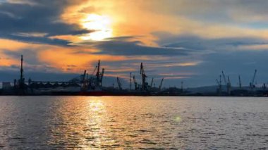 Gün batımında bulutlu gökyüzüne karşı çoklu konteynır vinçlerinin gümbürtüleri. Su manzarasından modern limana bak.