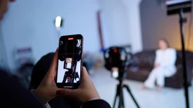 Beyaz takım elbiseli bir kadının bulanık arka planda otururken fotoğraflarını çeken kadın elleri telefonu tutuyor. Kamera arkası fotoğraf stüdyosu görüntüleri.