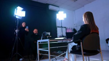 Kadın blogcu masanın önünde kameranın önünde oturuyor. Düşük açı. Kadın ve erkek arka plandaki stüdyoda video çekiyor..
