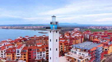 Deniz feneri kulesi, sahil kasabasının arka planına karşı. Beyaz bir deniz feneri kulesinin arka planında binalar ve bulutlu bir gökyüzünün altında deniz olan orta büyüklükte bir görüntüsü..