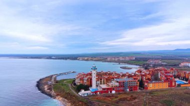 Deniz feneri ve kırmızı binaları olan sahil kasabasının havadan görünüşü. Beyaz bir deniz feneri, kırmızı bir mimari ve bulutlu bir gökyüzünün altındaki kıyı şeridini gösteren yüksek açılı bir deniz kenti manzarası..