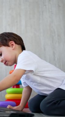 Küçük oğlan dizüstü bilgisayarın önünde yerde oturuyor. Yürüyen çocuk klavyedeki tuşlara basıyor. Düşük açı. Dikey video