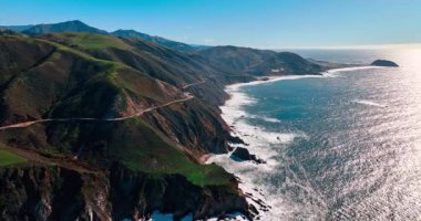 Big Sur kıyısı boyunca kıvrılan 1. otoyolun panoramik hava manzarası. Virajlı yol, dik uçurumlar ve mavi okyanus ufkunun geniş açılı görüntüsü.