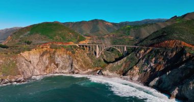 Bixby Creek Köprüsü hava manzaralı, okyanus dalgaları ve dağlar. Big Sur 'da kumlu bir sahil üzerinde kanyon boyunca uzanan tarihi beton kemer köprüsü
