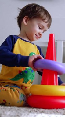 Tatlı erkek bebek oyuncak piramidinin yanında yere oturuyor. Çocuk oyuncağı neşeyle monte ediyor. Düşük açı. Dikey video