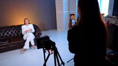 Akıllı telefon çekim röportajının omuzlarının üstünden. Beyaz takım elbiseli bir kadın profesyonel stüdyoda kameralı cep telefonu kullanıyor..