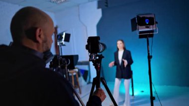 Kamera ekranı, bayan sunucunun mavi stüdyo arka planında durduğunu gösteriyor. Kamera ekranındaki kameramanın arkasından beyaz elbiseli kadını profesyonel ışıklandırma ekipmanıyla görüntüle.