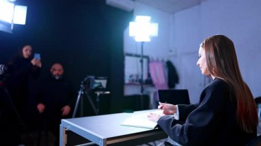 Video çekimi sırasında masasında siyah ceketli bir kadın sunucu. Stüdyoda, arka planda kamera ekibi ve profesyonel ışıklandırma ekipmanıyla birlikte masa başında oturan bir kadın..