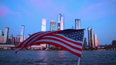 Su manzarası ve şehir silueti üzerinde dalgalanan Amerikan bayrağı. Arkaplanda gün ışığında gökdelenler.