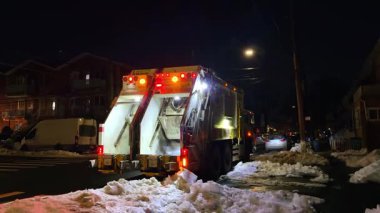 New York, ABD, 9 Şubat 2026: Geceleri karlı iskan caddesinde çalışan temizlik kamyonu. Kış boyunca şehir sokaklarında çöp toplayan parlak arka lambalı bir çöp kamyonu..