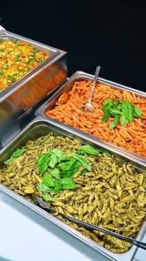 Makarna ve pirinç sebze tabağı ile büfe masası. Pesto penne, domatesli makarna ve karışık sebzeli sarı pirinç içeren açık büfe kuyruğu..