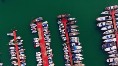 Limandaki düzenli yat sıralarının hava görüntüsü. Koyu yeşil deniz suyuna kenetlenmiş beyaz tekneleri olan birden fazla kırmızı iskele dikey görünümü.