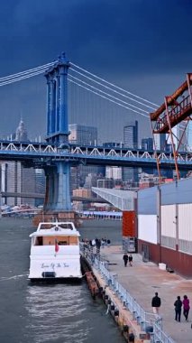 New York, ABD, 25 Ağustos 2025, Manhattan Köprüsü Doğu Nehri üzerinde iskeleye yanaşmış bir tekne. Nehir üzerinde mavi çelik asma köprü, rıhtımda beyaz yolcu teknesi ve şehir binaları var..