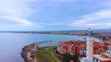 Beyaz deniz feneriyle deniz kenarındaki tatil köyünün yüksek açılı manzarası. Sahil köyünde kırmızı çatılar ve durgun turkuaz deniz kenarındaki uzun beyaz deniz feneri kulesi.