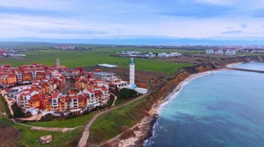 Kırmızı çatısı ve uzun deniz feneri olan sahil beldesi. Akdeniz tarzı apartman kompleksi ve deniz kıyısındaki beyaz deniz fenerinin havadan çekilmiş görüntüleri..