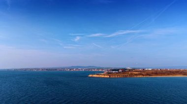 Geniş kıyı şeridi manzarası belirgin uçurumu ve mavi gökyüzü zıtlıkları. Kayalık bir nokta, kumlu plaj ve beyaz çizgili mavi gökyüzü içeren uzak bir kıyı şeridi manzarası.