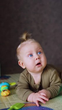 Sarışın erkek bebek oyuncakların arasında yerde yatıyor. Tatlı bebek kameraya doğru emeklemeye çalışıyor. Dikey video