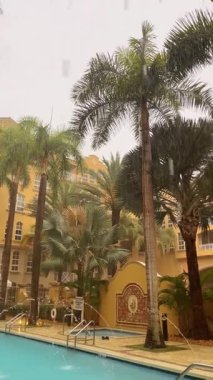 Florida 'da açık havuza ve palmiye ağaçlarına yağmur yağıyor. Sarı Akdeniz tarzı bina ve tropikal bitki örtüsü bulunan havuz üzerinde yoğun yağmur damlacıkları.