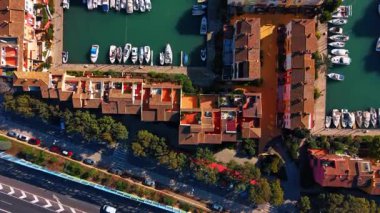 Rıhtımda daireler ve demirli tekneler. Yakındaki bir yol ve yeşillik olan bir kanalda terracotta çatıları ve yatları gösteren detaylı hava manzarası var..