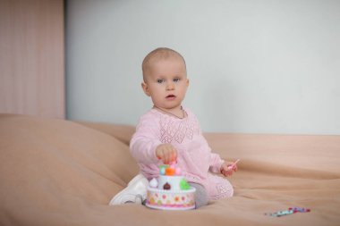 Yatağın üzerinde pembe elbiseli bir yaşında bir bebek oturuyor.