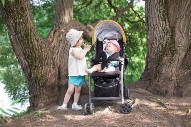 Yazın parkta iki küçük kız kardeş. Pembe Panama şapkalı en büyüğü. Küçük olan sarı tekerlekli sandalyede oturuyor..