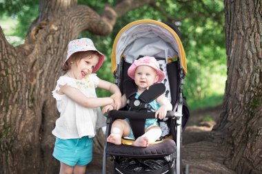 Yazın parkta iki küçük kız kardeş. Pembe Panama şapkalı en büyüğü. Küçük olan sarı tekerlekli sandalyede oturuyor..