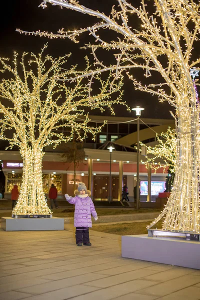 A European girl in a lilac down jacket stands among the festive trees