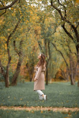 Pembe ceketli ve kot pantolonlu beyaz bir kız sonbahar parkında yürüyor.