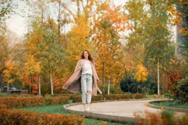 Pembe ceketli ve kot pantolonlu beyaz bir kız sonbahar parkında dans ediyor.