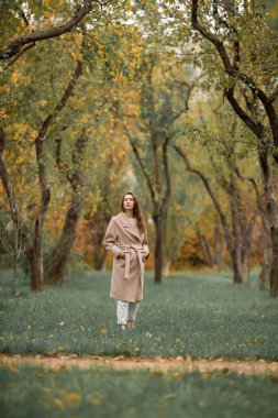 Pembe ceketli ve delikleri olan mavi kot pantolonlu bir kız sonbahar parkında yürüyor.